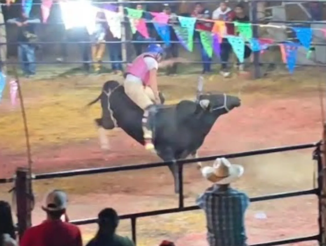 Jinete montando un toro en el ruedo, entre banderines de papel picado